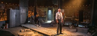 An actor portraying Jim McLean stands centre stage in a theatre set resembling a construction site and office, with another actor seated in the background, amidst a scene from the production 'Smile.'"