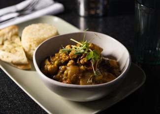 A bowl of lentil and vegetable curry garnished with fresh herbs, accompanied by warm pita bread on a sleek serving platter, set against a dark tabletop.