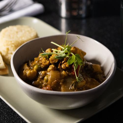 A bowl of lentil and vegetable curry garnished with fresh herbs, accompanied by warm pita bread on a sleek serving platter, set against a dark tabletop.