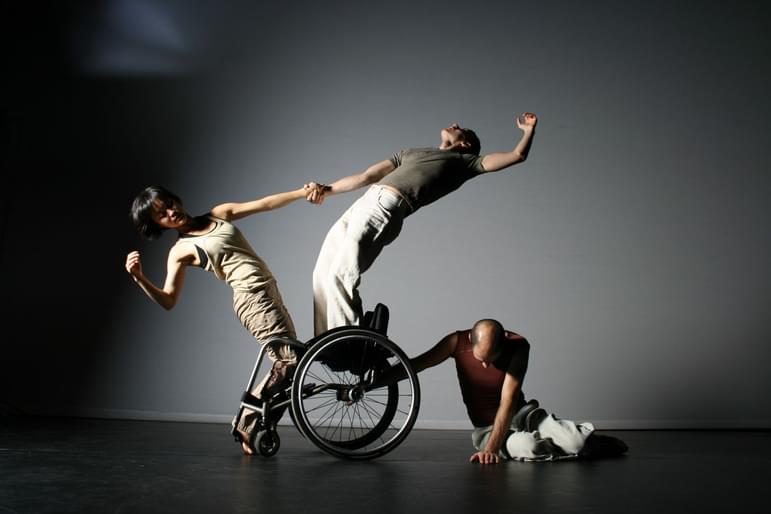 3 dancers stand on or hold onto a wheelchair in different stretched poses