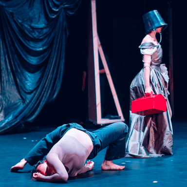 A dancer contorts backwards on the floor in front of another standing wearing a baroque dress, a bucket on their head and carrying a red toolbox