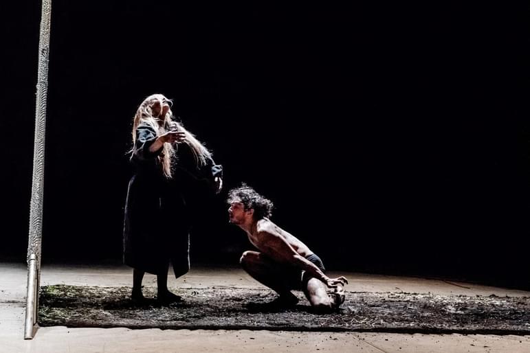 A woman stares towards the sky standing on a muddy patch of ground on a stage as a man crouches shirtless at her feet