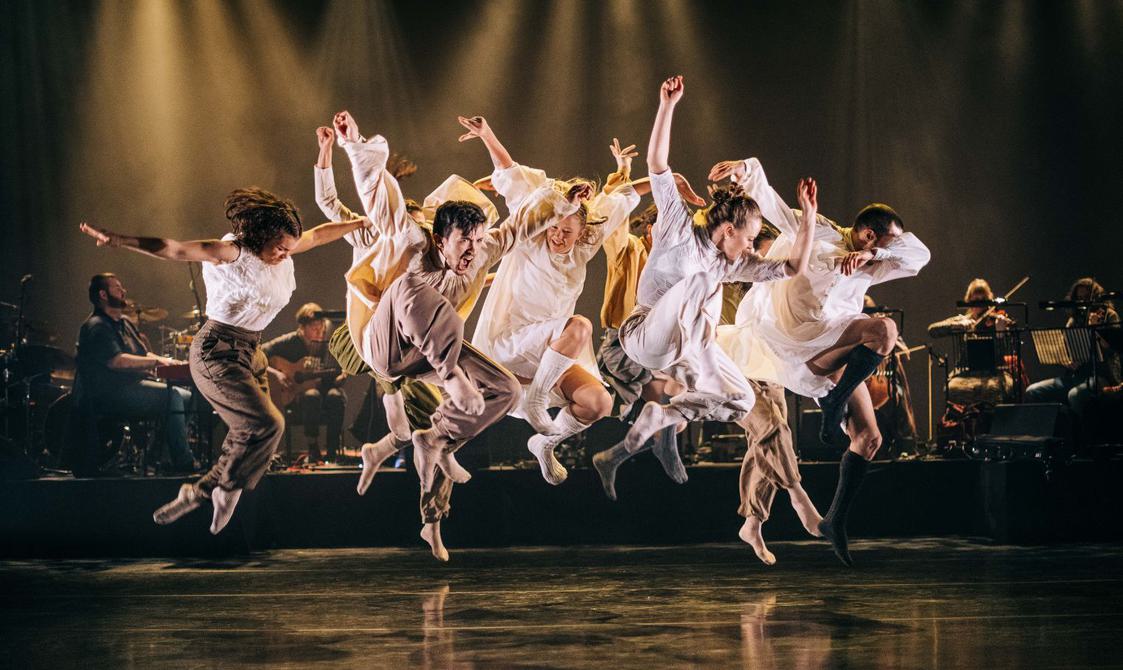 A group of dancers leap with energy above a black stage lit in golden light, wearing loose white, cream and brown clothing