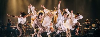 A group of dancers leap with energy above a black stage lit in golden light, wearing loose white, cream and brown clothing