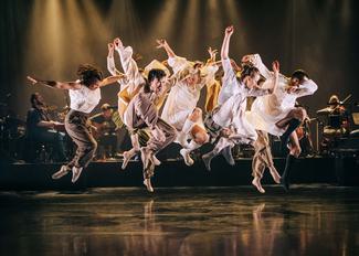 A group of dancers leap with energy above a black stage lit in golden light, wearing loose white, cream and brown clothing