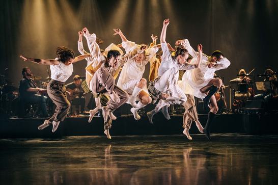 A group of dancers leap with energy above a black stage lit in golden light, wearing loose white, cream and brown clothing