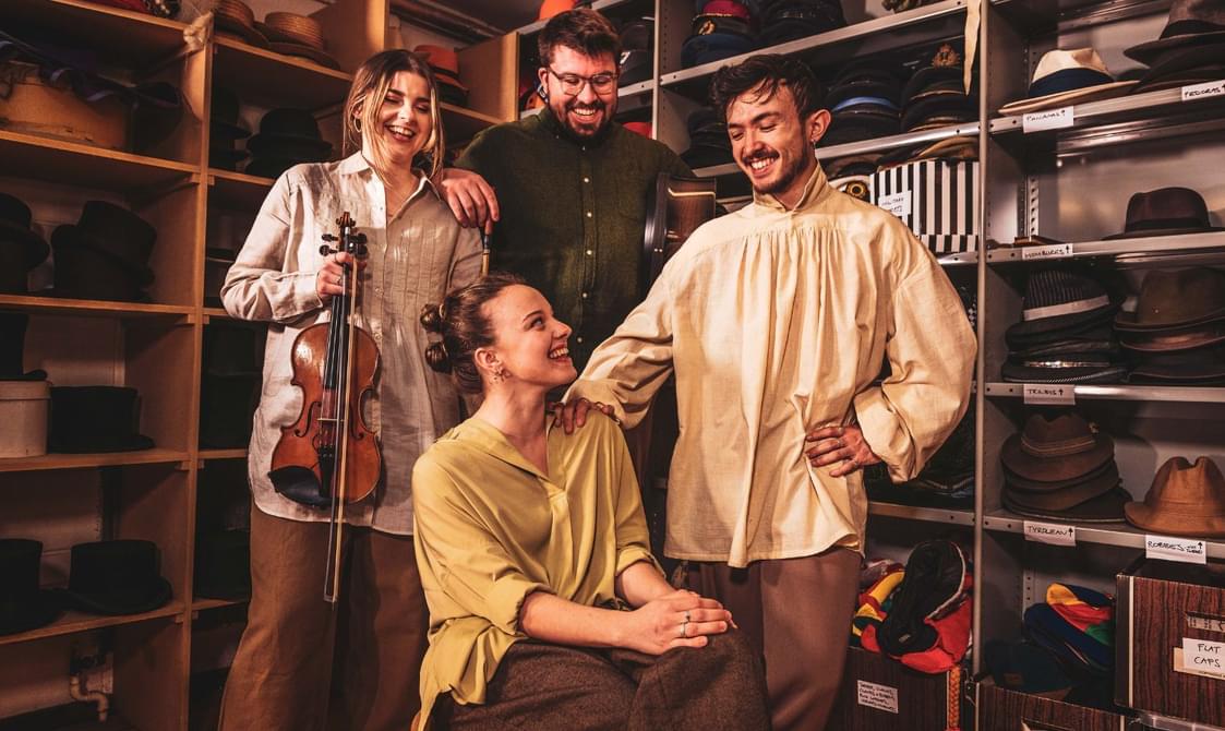 A girl holding a violin stands next to a bearded man in a green shirt and a man with a moustache and dark hair in a loose cream shirt all looking down laughing at a seated woman in a pale green shirt surrounded by hats on shelves