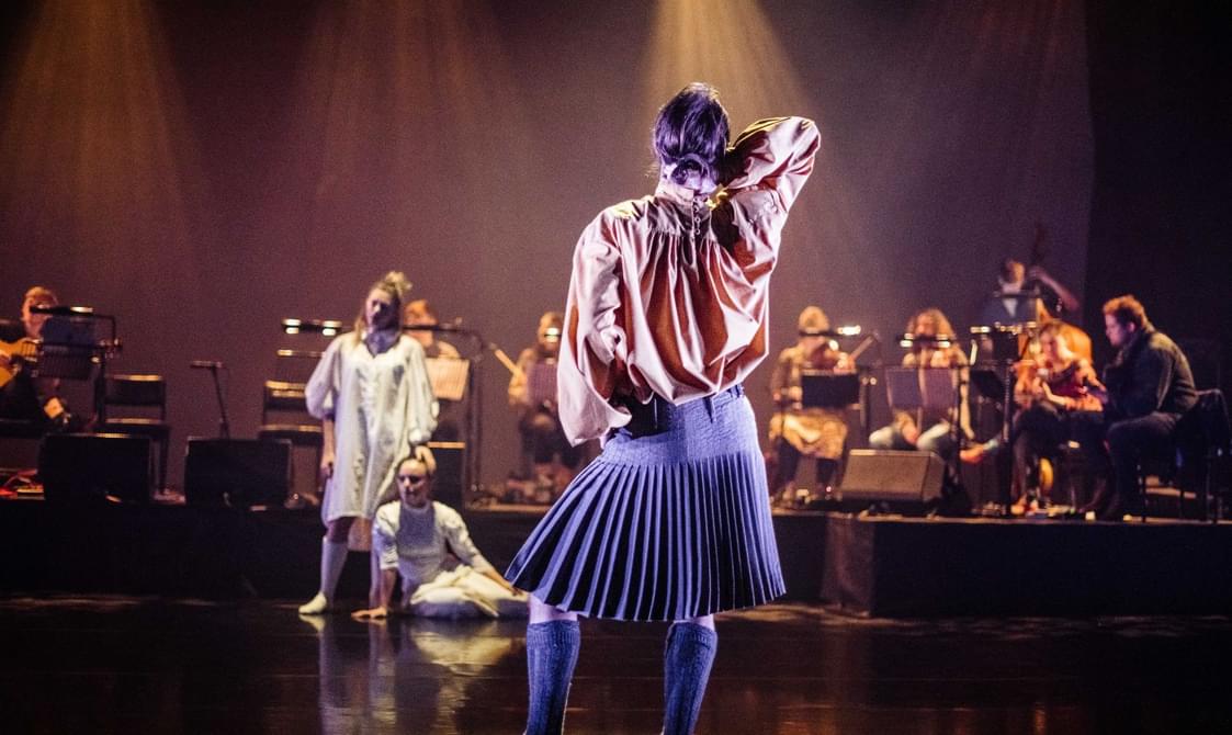 A man stands in spotlight facing the back of a stage in a cream loose shirt and a grey kilt and socks with a band behind him
