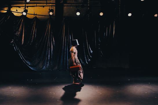 A person stands with a bucket on their head in front of a black curtain holding a red toolbox