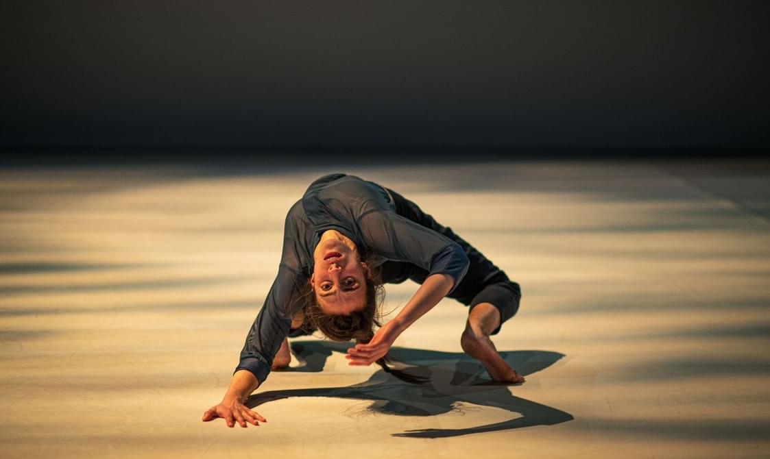 A female dancer with dark hair and grey loose clothing crouches in a back bend in a contorted pose