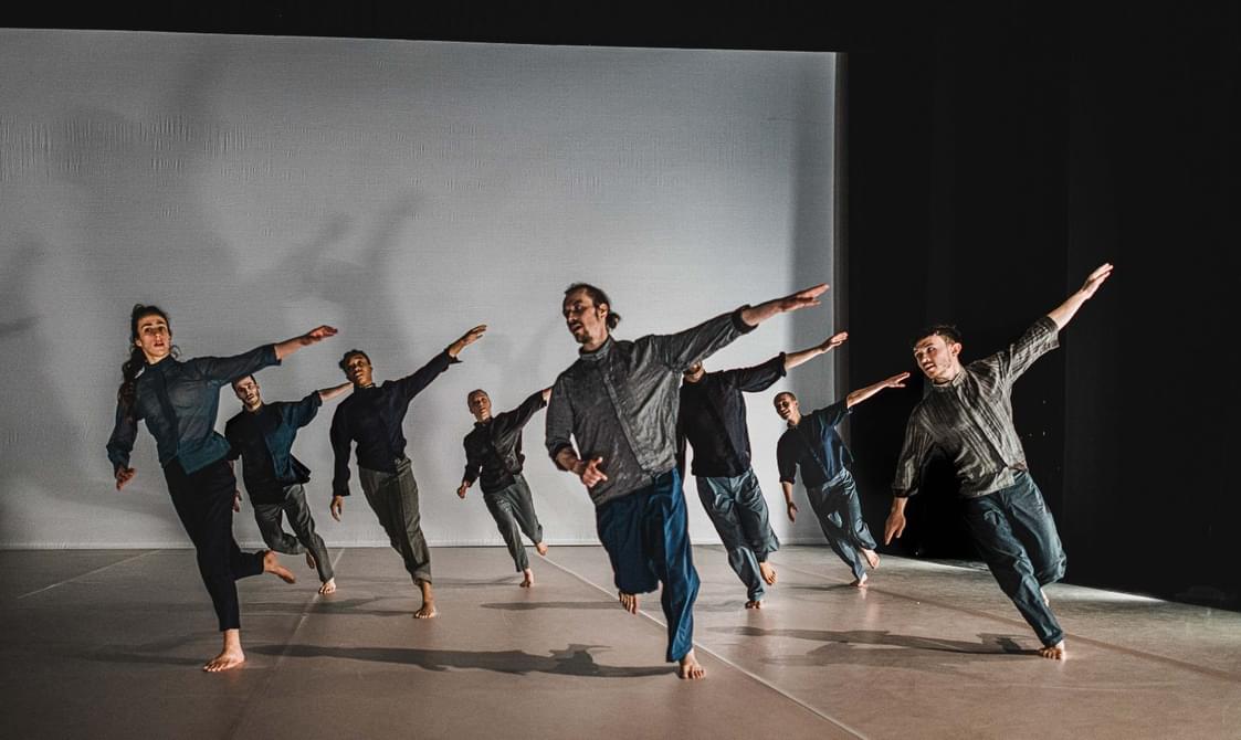 A group of dancers run in formation with left arms outstretched on a grey floor