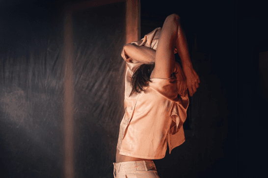 A woman contorts her arms around her head so her face isn't visible wearing a light pink short-sleeved shirt and cream trousers on a black background