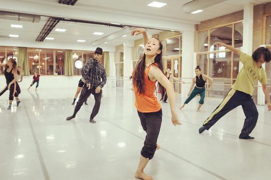 A group of dancers practicing in a brightly lit dance studio with mirrors along one wall. One dancer in the foreground is wearing an orange top and black pants, visibly captured in an expressive dance move with her arm gracefully arched overhead.