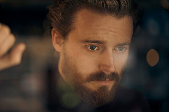 A man with beard and moustache leans against a window staring forward just past the camera with a sombre look