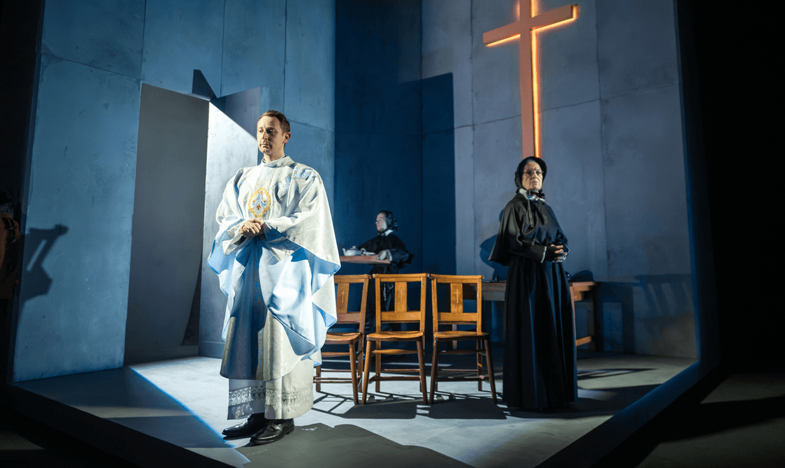 The Doubt: A Parable cast stand on a dark stage lit by a golden cross