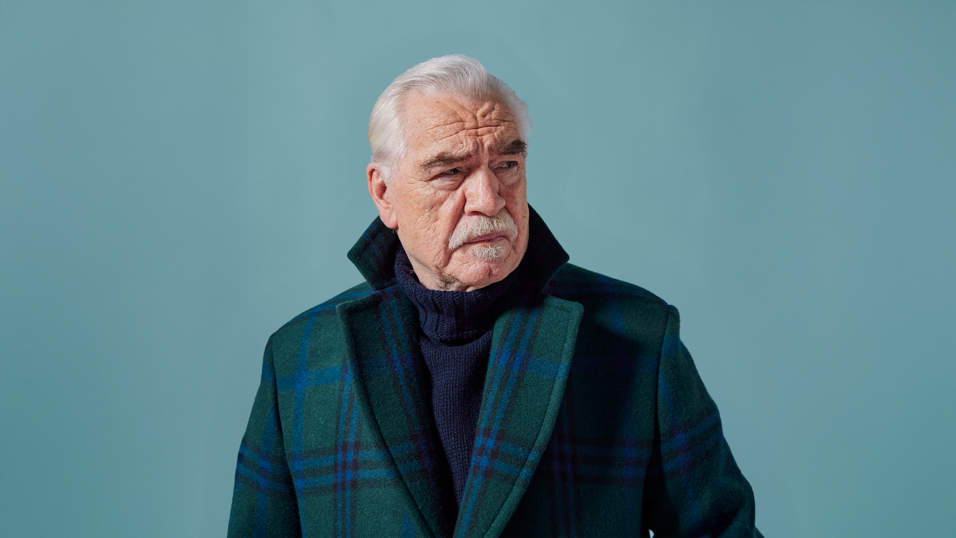 Actor Brian Cox looks into the distance wearing a green and blue tartan coat against a blue background