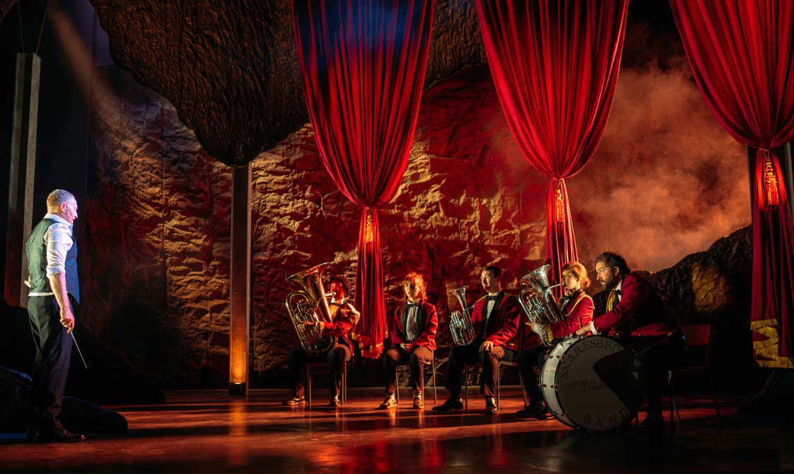 KELI production photo of a group od people in red coats holding brass instruments on a stage