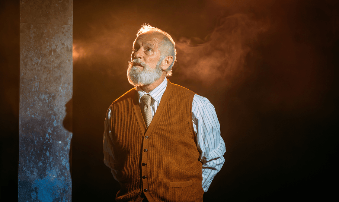 An old man with a well-kempt grey beard lit in gold wearing a gold tie, pinstriped shirt and brown waistcoat looks to the sky