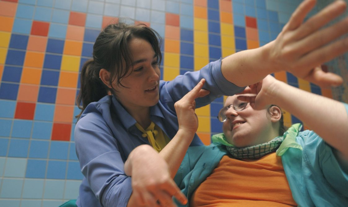 Two woman dance in front of a tiled colourful wall