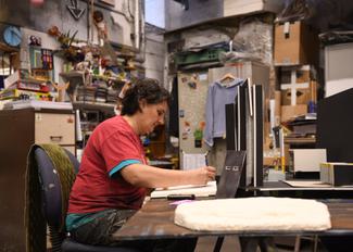 A woman is sitting at a desk, working on a model set for a theatrical production. She is in a workshop, surrounded by boxes and random objects.