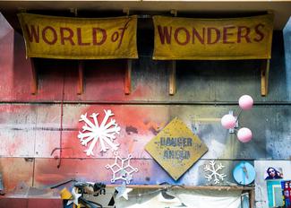 A wall in a theatrical paintshop, featuring a worn and artistic backdrop painted with streaks of red and blue. Two tattered yellow banners proclaiming 'WORLD of WONDERS' drape from above, complementing the assortment of objects below, which includes a bold white snowflake decoration, a yellow 'DANGER AHEAD' sign, pink orbs, and snippets of imagery and cut-outs.