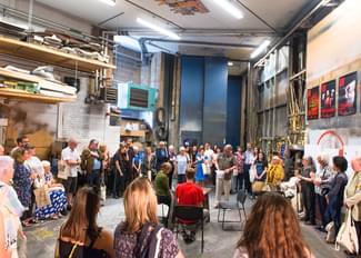 A group of people are gathered in a circle in a large workshop. At the centre of the circle are three actors rehearsing a scene for the crowd.