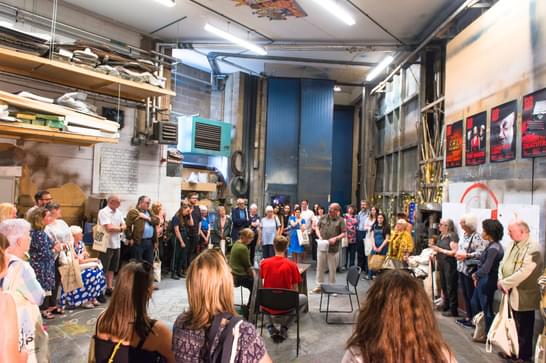 A group of people are gathered in a circle in a large workshop. At the centre of the circle are three actors rehearsing a scene for the crowd.