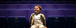 A cheerful small boy sits in an empty theatre auditorium
