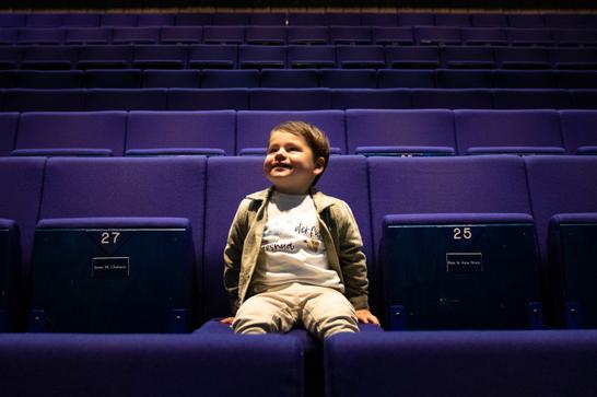 A cheerful small boy sits in an empty theatre auditorium