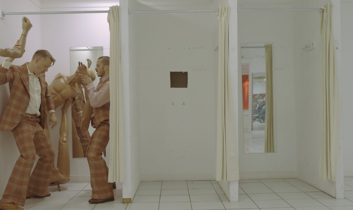 Two men dance wearing checked suits in the left cubicle of three in a shop changing room