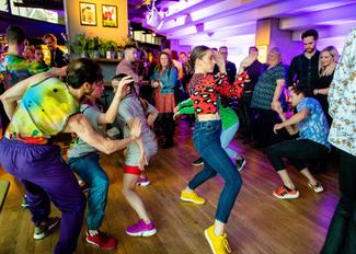 A vibrant dance scene with a group of people in casual attire, energetically dancing and enjoying themselves at an indoor venue with colorful lighting.