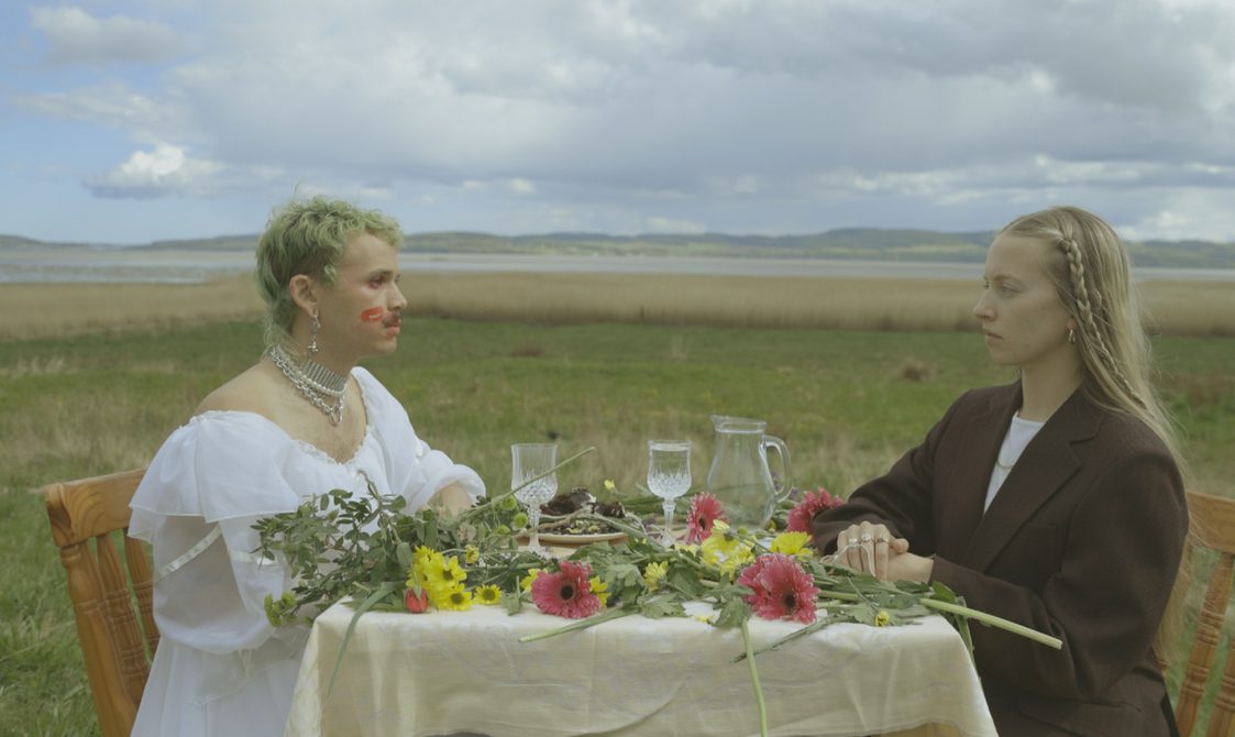 Two people sit at a table covered in flowers in front of a field