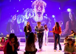 A production shot for the show Optimism which shows a group of young people on stage with one performer shouting into a camera. The image from the camera is being projected onto the back wall of the stage.