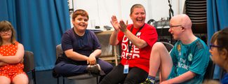 A group of children and an adult enjoying an interactive session indoors. One child in a red polka dot dress sits on the left, next to a smiling boy in a navy t-shirt. An adult in a red 'Dive Into Books' t-shirt and shorts is clapping with joy, seated next to a child in a teal Real Madrid jersey and glasses. They are all seated on chairs in a semi-circle, with a blue curtain and music equipment in the background.