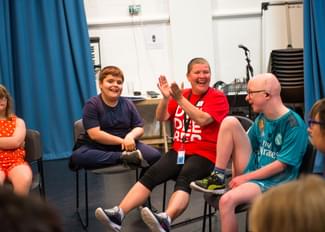 A group of children and an adult enjoying an interactive session indoors. One child in a red polka dot dress sits on the left, next to a smiling boy in a navy t-shirt. An adult in a red 'Dive Into Books' t-shirt and shorts is clapping with joy, seated next to a child in a teal Real Madrid jersey and glasses. They are all seated on chairs in a semi-circle, with a blue curtain and music equipment in the background.