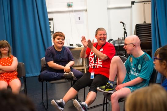 A group of children and an adult enjoying an interactive session indoors. One child in a red polka dot dress sits on the left, next to a smiling boy in a navy t-shirt. An adult in a red 'Dive Into Books' t-shirt and shorts is clapping with joy, seated next to a child in a teal Real Madrid jersey and glasses. They are all seated on chairs in a semi-circle, with a blue curtain and music equipment in the background.