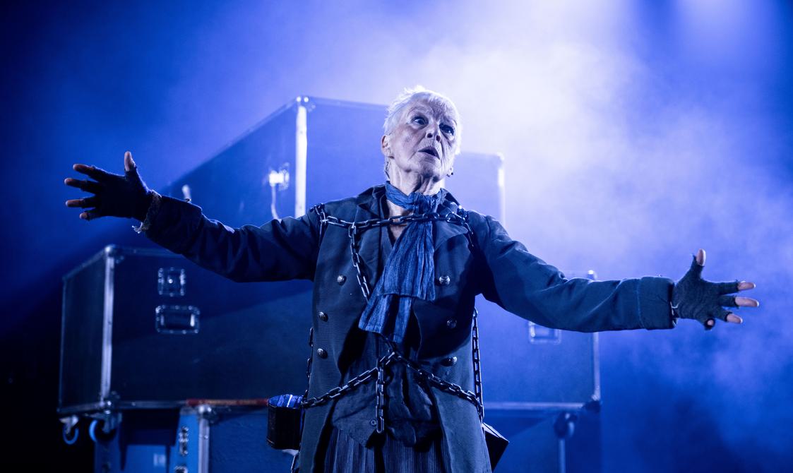 A woman with white short hair stands with arms outstretched wearing a blue cravat and dark overcoat, lit dramatically with blue and white light and haze in front of a stack of flight cases
