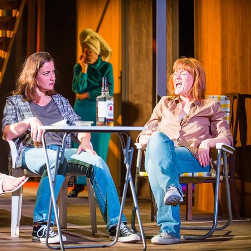 A image from a theatrical production of 'August: Osage County' showing three actresses on stage. From left to right, the first actress is seated on a folding chair, holding a cigarette, with a pensive expression. The second, in the center, is seated with her hands clasped, looking intently at the third actress. The actress on the right is holding a bottle of liquor and gesturing with her other hand, portraying a scene of dialogue. The backdrop is minimally designed to resemble a porch setting.