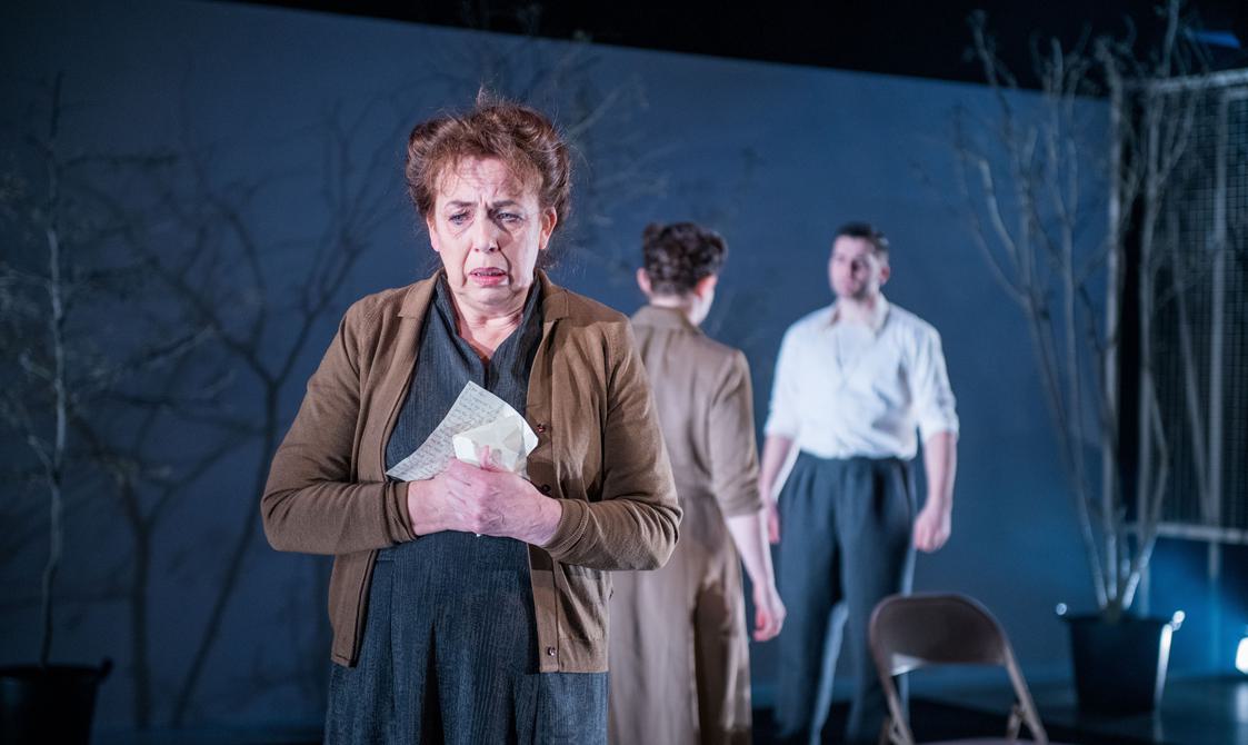A promotional image from a theatrical production of 'All My Sons' showing an actress in the foreground. She is clutching a handwritten letter in her hands with a shocked yet sad expression. In the background, an actress and actor stand facing each other as in conversation.