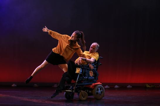 A dance theatre performance featuring a dancer in a yellow top and black shorts, with a person in a yellow shirt seated in a motorized wheelchair on a dimly lit stage.