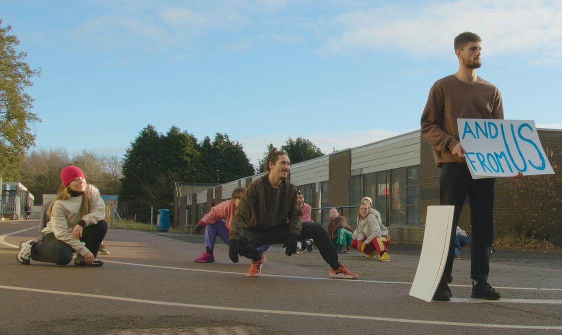 A scene from one performance of 'Present' featuring individuals in casual wear actively engaging in a street performance. One participant is standing, holding a placard that reads 'AND US FROM US', while others are captured in various crouched positions, conveying a sense of movement.