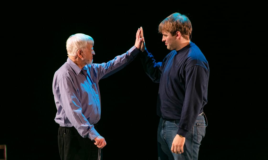 An older man with white hair wearing a purple shirt and a younger man in a dark blue sweater giving a high-five on a stage with a black background.