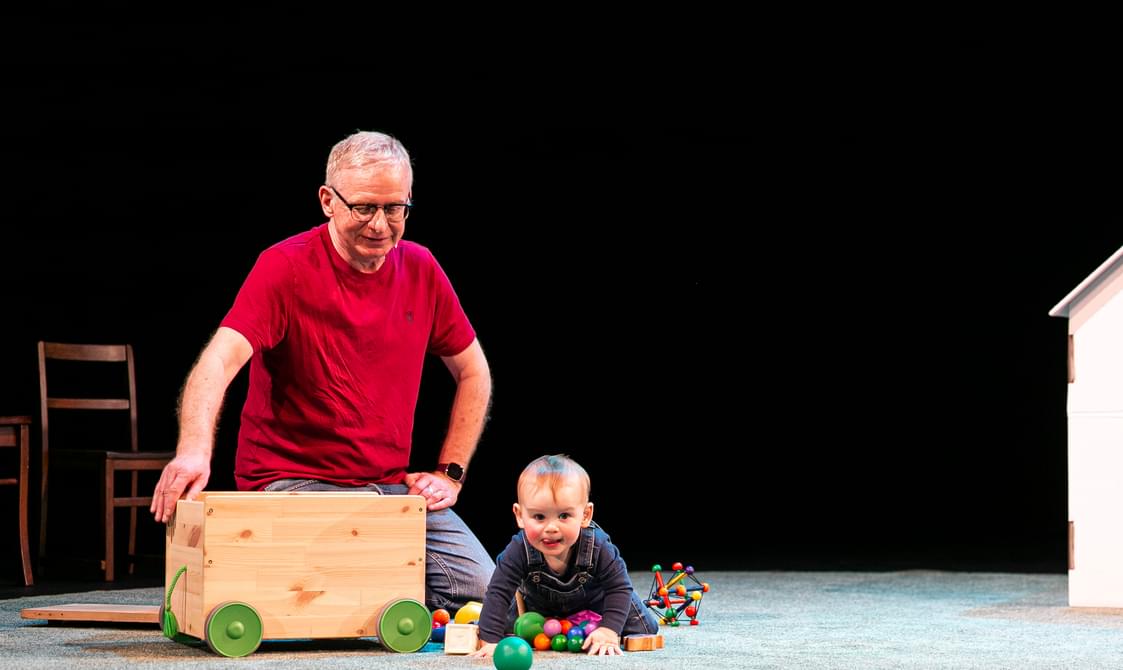 A smiling older adult in a red shirt and glasses is sitting behind a large wooden toy box on wheels. In front of him, a happy infant is playing with colourful balls on a stage set up to resemble a home, with a small white door frame on the right side.