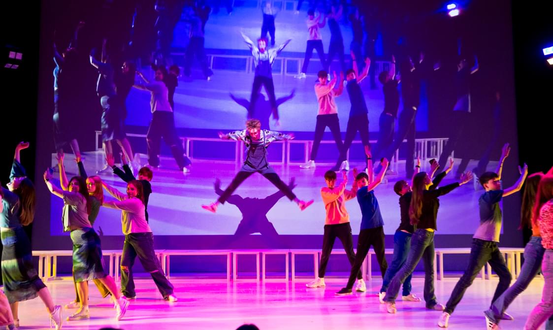 A dynamic stage performance featuring a central performer in mid-leap with arms extended, surrounded by a vibrant ensemble of performers in colourful costumes, against a backdrop of a projected live image of what is happening on stage.