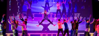 A dynamic stage performance featuring a central performer in mid-leap with arms extended, surrounded by a vibrant ensemble of performers in colourful costumes, against a backdrop of a projected live image of what is happening on stage.