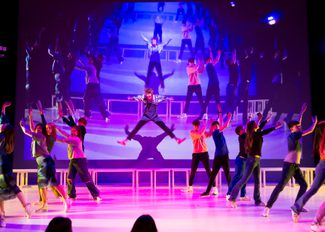 A dynamic stage performance featuring a central performer in mid-leap with arms extended, surrounded by a vibrant ensemble of performers in colourful costumes, against a backdrop of a projected live image of what is happening on stage.