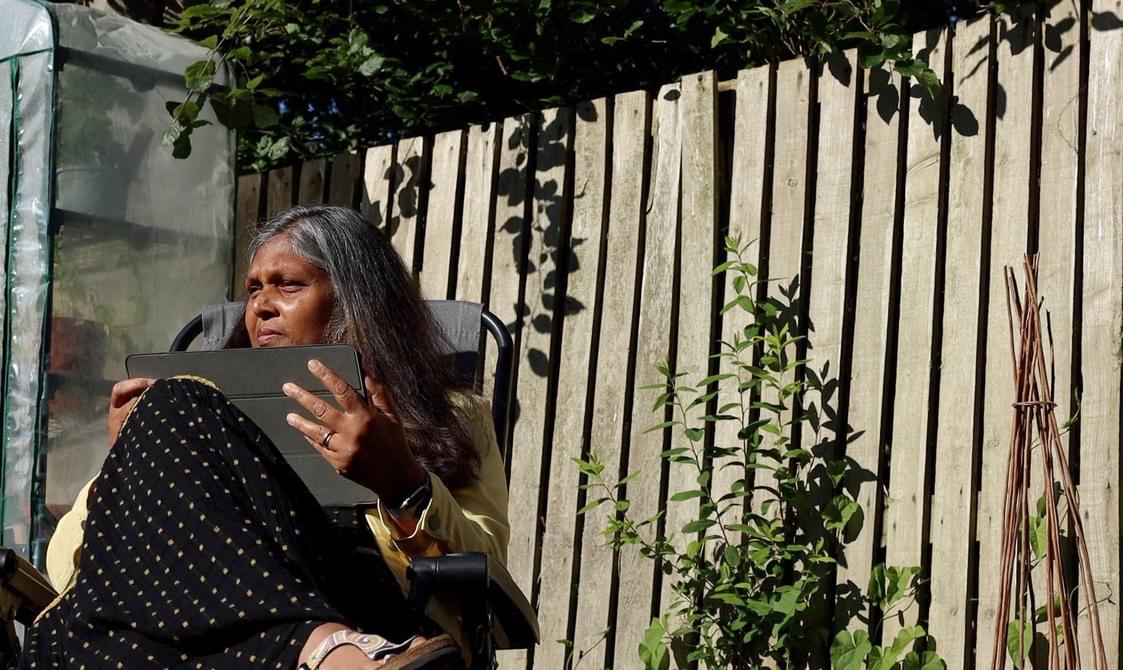 A woman with grey hair, wearing a yellow shirt and patterned shawl, is seated in. She is holding an ipad in her hands. The background features a wooden fence with some greenery under a bright sunny sky.