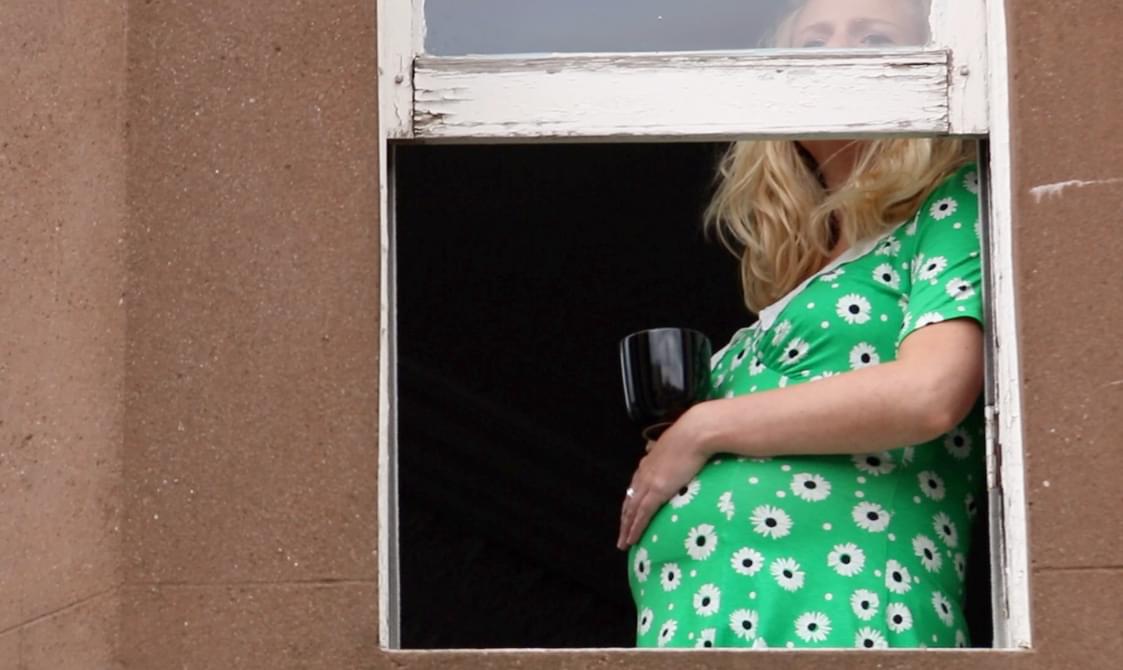 A pregnant woman in a green dress with white polka dots is standing by an open window, partially visible from the waist up. The window frame is white and chipped, set in a brown wall.