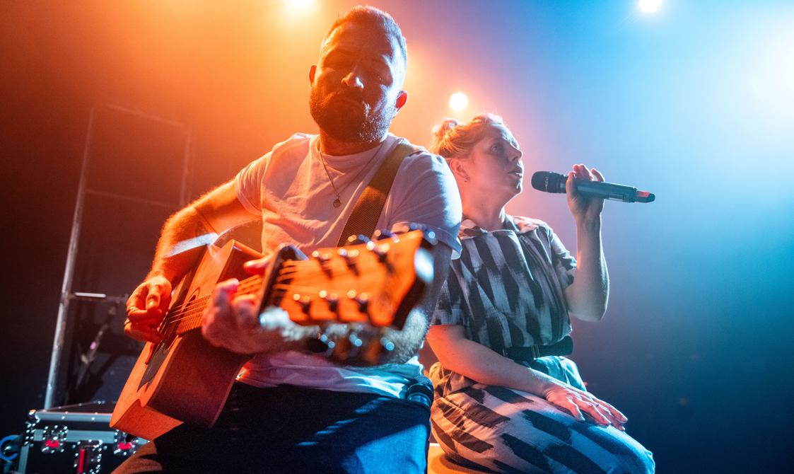 A man wearing a white tshirt and jeans holding a guitar sits next to a woman in a black and white jumpsuit holding a microphone on two flight cases