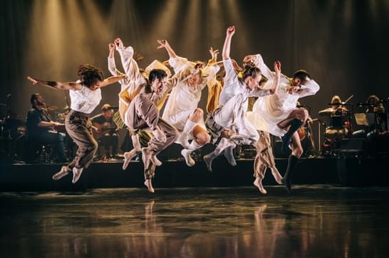 A group of dancers performing on stage with a live orchestra in the background during the 'Moving Cloud' dance production.